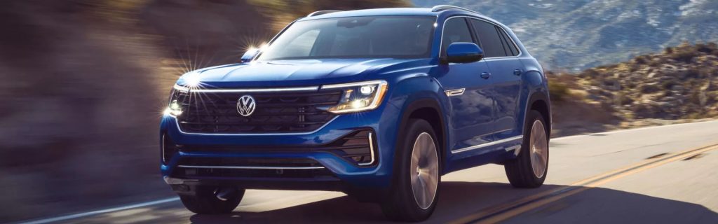 Blue 2026 Volkswagen Atlas Cross Sport driving on a scenic mountain road with sunlight reflecting off the front grille.